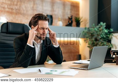 Headache, Migraine. Fatigue Caucasian Businessman, Office Employee, Sits At A Workplace In A Modern 