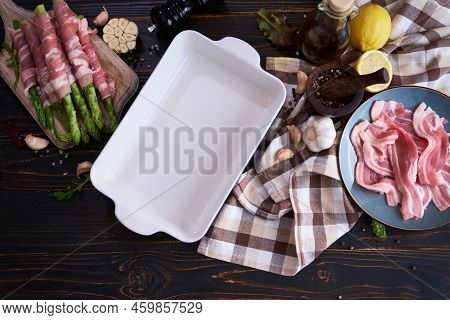 Ceramic Baking Dish For Cooking Aspargus Covered With Bacon On Wooden Kitchen Table