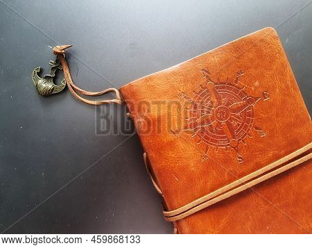 A Notebook With A Brown Leather Cover With A Compass Motif And Anchor Strap.