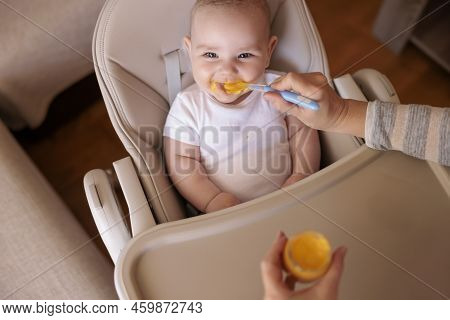 High Angle View Of Mother Feeding Cheerful Baby Boy With Porridge, Baby Sitting In High Chair All Me