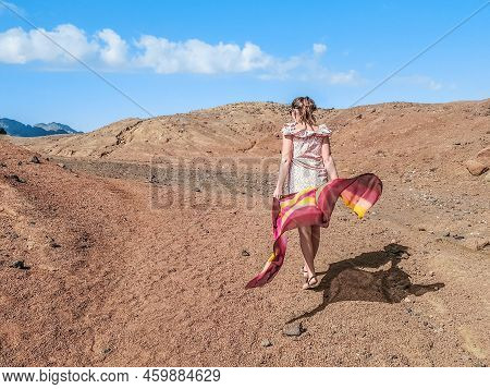 A Lonely Adult Girl With A Light Scarf In Her Hands Walks Through The Desert - View From The Back. A