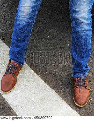 The Lower Of The Legs Of A Standing Man Wearing Blue Denim Jeans With Brown Leather Shoes.