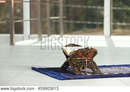 Rehal With Open Quran And Misbaha On Muslim Prayer Rug Near Window Indoors, Space For Text