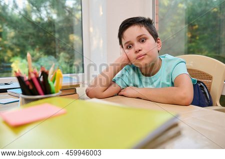 Tired And Bored Schoolboy, Primary Student In Online School, Sitting At Desk With School Supplies An