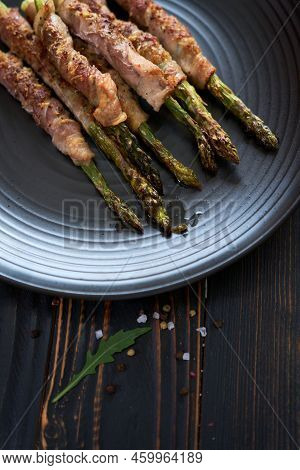 Baked Aspargus Covered With Bacon On A Black Ceramic Plate On Wooden Kitchen Table