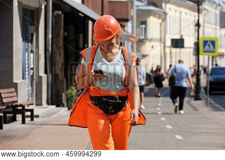 Moscow, Russia - August 2022: Woman Building Worker In Orange Uniform And Helmet Walking With Smartp