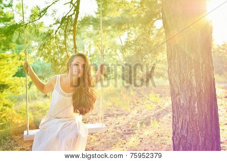Young woman is swinging on a swing in summer pine forest. Image toned and noise added. High key.