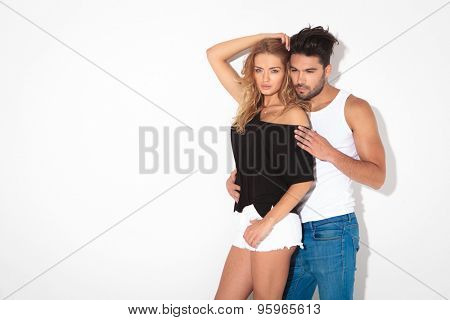 Fashion couple leaning on a white wall, the man is embracing the woman from behind.