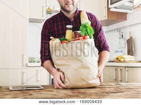 Man holding a paper bag full of healthy food on a wooden table at home