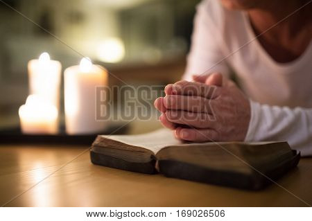 Unrecognizable senior woman lying on the floor praying with hands clasped together on her Bible. Burning candles next to her. Close up.
