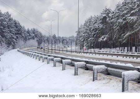 Part of the motorway with cars traffic barriers lampposts and pine forest covered snow on both sides of road after snowfall in cloudy day