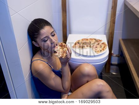 young sad and depressed bulimic woman feeling sick guilty sitting at the floor of the toilet leaning on WC eating pizza in nutrition disorder bulimia concept