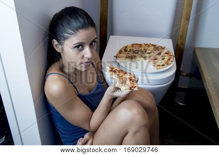 young sad and depressed bulimic woman feeling sick guilty sitting at the floor of the toilet leaning on WC eating pizza in nutrition disorder bulimia concept