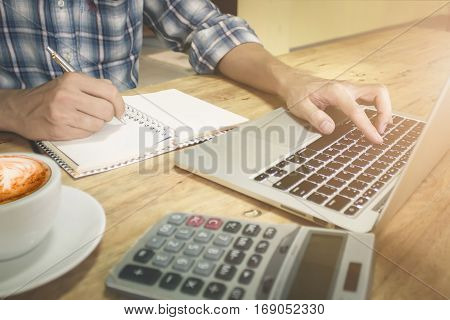 Business man using laptop computer with hand holding pen point notebook and coffee hot latte art. on wood desk. vitage tone.