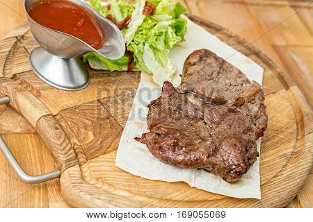 cooked steak on a pita a wooden board standing next to a gravy boat with ketchup and greens