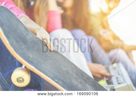 Young skaters friends listening music and chatting in city park - Young people having fun sitting outdoor - Youth and friendship concept - Focus on left girl hand - Warm cinematic filter