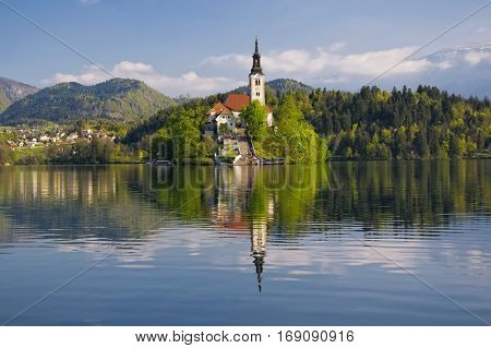 Island on Bled lake and pilgrimage church of the Assumption of Mary with mountain landscape background reflected in water at sunny day, Slovenia