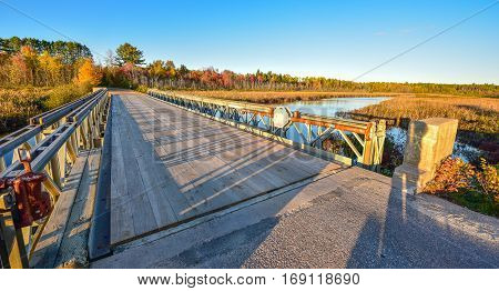 Wide angle view of clear blue sky, late afternoon sunshine, casts long shadows on a light steel and timber bridge, single point perspective over a lake in Ontario, Canada.
