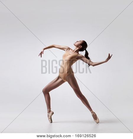 young beautiful ballet dancer in beige swimsuit posing on pointes on light grey studio background