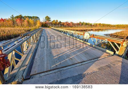 Wide angle view of clear blue sky, late afternoon sunshine, casts long shadows on a light steel and timber bridge, single point perspective over a lake in Ontario, Canada.