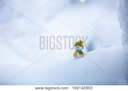 Nice dreaming snowman in green hat is standing on interesting textured snow near to the hill on cold winter day.