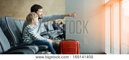 panorama of happy family of two father and son waiting together at the airport for plane departure with luggage father pointing with finger at something sun flare effect