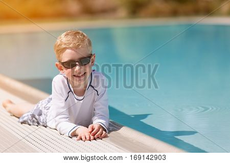 smiling cheerful boy in sunglasses lying by the pool in rashguard sun flare sun protection concept