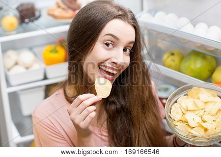 Young woman eating potato chips near the refrigerator full of healthy food