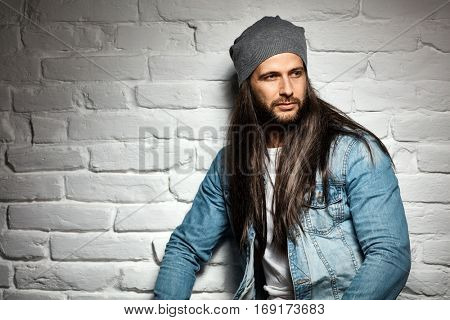 Portrait photo of rocker guy in sunglasses and woolly hat standing against brick wall.