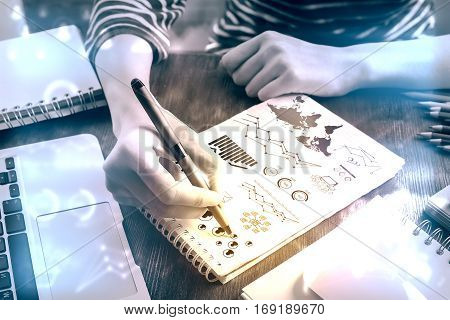 Top view of hands srawing business charts and diagrams on spiral notepad placed on wooden desktpop with laptop and other items. Filtrered image