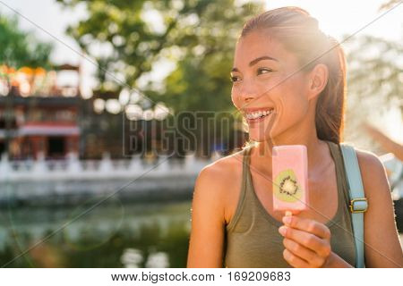 Happy asian tourist woman eating healthy enjoying frozen treat snack in summer park in Beijing hutong, near Houhai lake. Popular urban destination for romantic getaway in China.