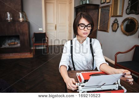 Authoress in glasses and white shirt sitting by the table and preparing typewriting