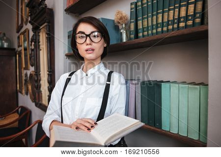 Authoress in glasses and white shirt standing near the bookshelf, holding book and looking at camera