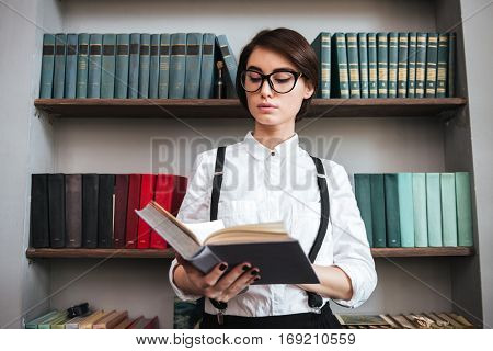 Authoress in glasses and white shirt reading book with bookshelf on background