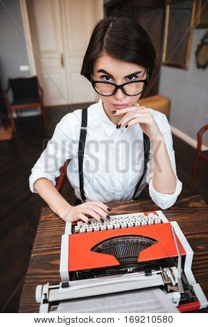 Vertical image of Concentrated Authoress in glasses and white shirt using typewriting and looking at camera