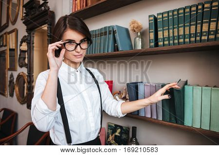 Pretty Authoress in glasses and white shirt selecting the book on bookshelf and looking at camera