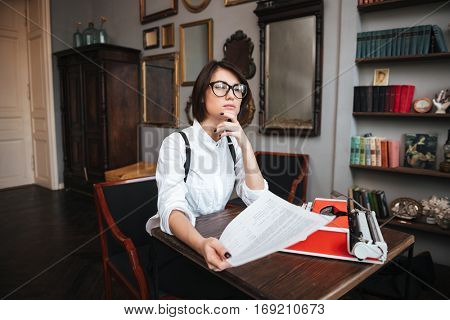 Authoress in glasses and white shirt sitting by the table with paper and typewriter and looking up
