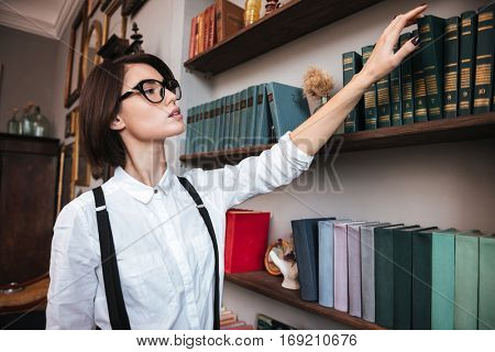 Authoress in glasses and white shirt selecting the book on bookshelf. Side view