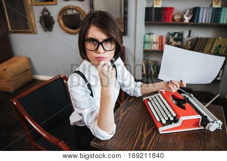 Side view of young authoress in glasses and white shirt sitting by the table with paper and typewriter and looking at camera. Top view