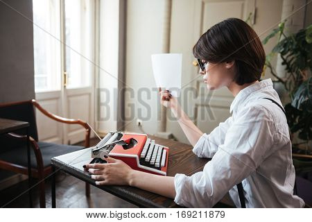 Side view of authoress in white shirt and glasses sitting by the table with paper and typewriter