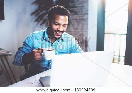 Smiling bearded African man using laptop home while drinking cup black coffee at the wooden table.Concept of young people working on mobile devices.Blurred window background