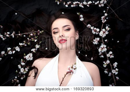 Spring beauty consept. Portrait of a fashionable model with curly brown hair sexy red lips lying on black background with spring trees blossoming branches and petals background. Studio portrait shot of gorgeous woman in white dress with perfect make-up se