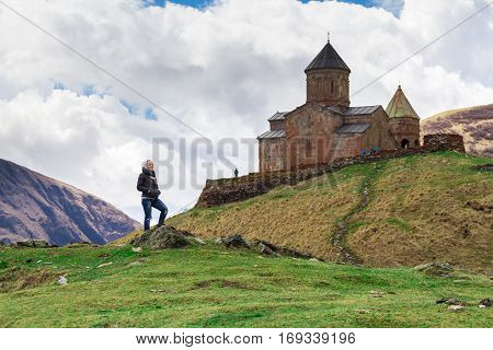 Female traveler on a background of Gergeti Trinity Church Or Tsminda Sameba in the morning mist near village Stepantsminda, Georgia. Mount Kazbegi. Spring Or Summer Season.