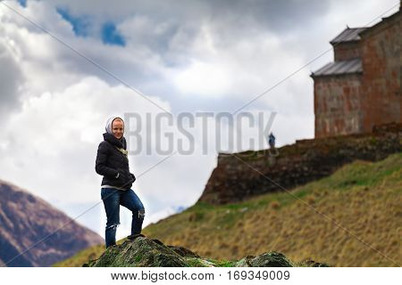 Female traveler on a background of Gergeti Trinity Church Or Tsminda Sameba in the morning mist near village Stepantsminda, Georgia. Mount Kazbegi. Spring Or Summer Season.