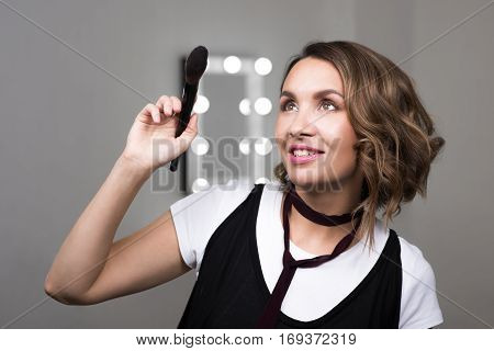 Portrait of a beautiful woman holding a makeup brush powder and looking up. Mirror with lights on background