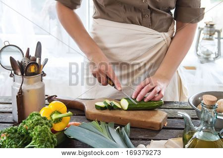 Woman slice zucchini on wooden table near window. Rustic style.