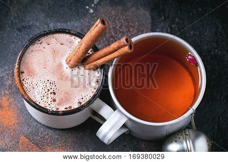 Two vintage mugs with tea and hot chocolate, served with dry tea, rose buds, chopped chocolate and cinnamon sticks over black background. Top view.