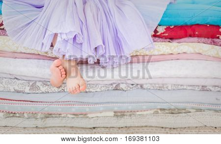 The fairy tale the princess on a pea. Little girl in lilac tatu skirt sitting on the high bed. Bare foot if the girl. Legs of a little girl sitting on a pile of colorful mattresses.