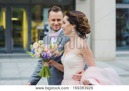 Happy bride and groom walking. Luxury bride holding a flying pink wedding dress and walking. Beautiful bride in a luxurious dress. Groom hugs bride walking on the street.