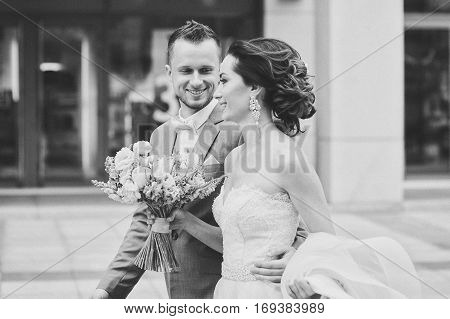 Happy bride and groom walking. Luxury bride holding a flying pink wedding dress and walking. Beautiful bride in a luxurious dress. Groom hugs bride walking on the street. black and white photo